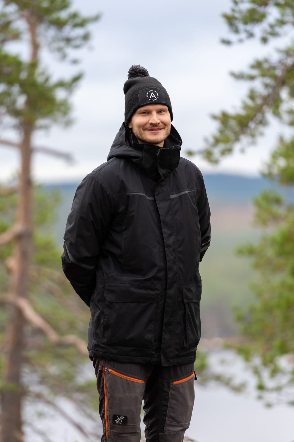 Esa Inget standing among pine trees above a lake, in an Aihki Adventures beanie.