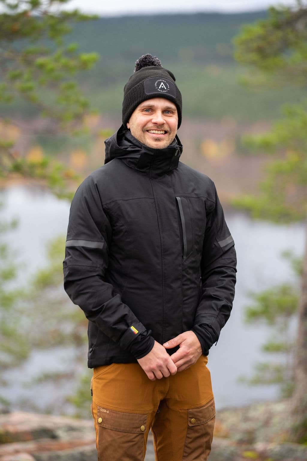 Erkka Gustafsson standing on a hill above a lake, smiling, in an Aihki Adventures beanie.