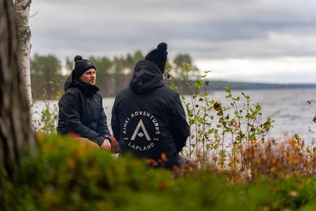 Erkka and Esa sitting by a lake in autumn, with an Aihki Adventures logo on the back of Esa’s jacket.