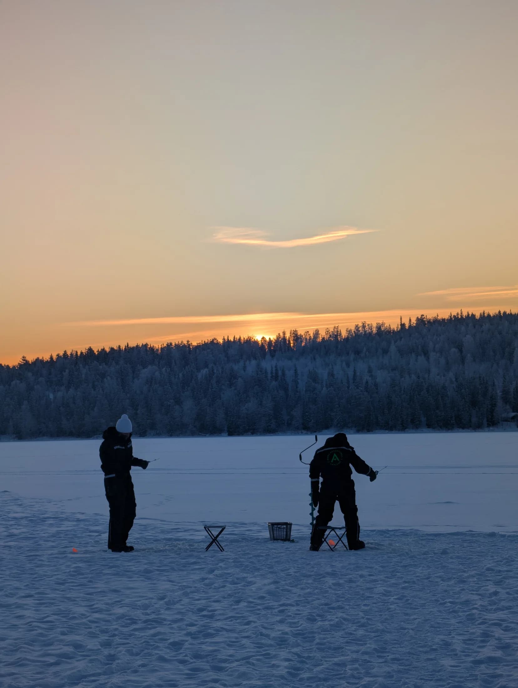 Two guests fishing on the frozen lake as the sun sets behind the forest